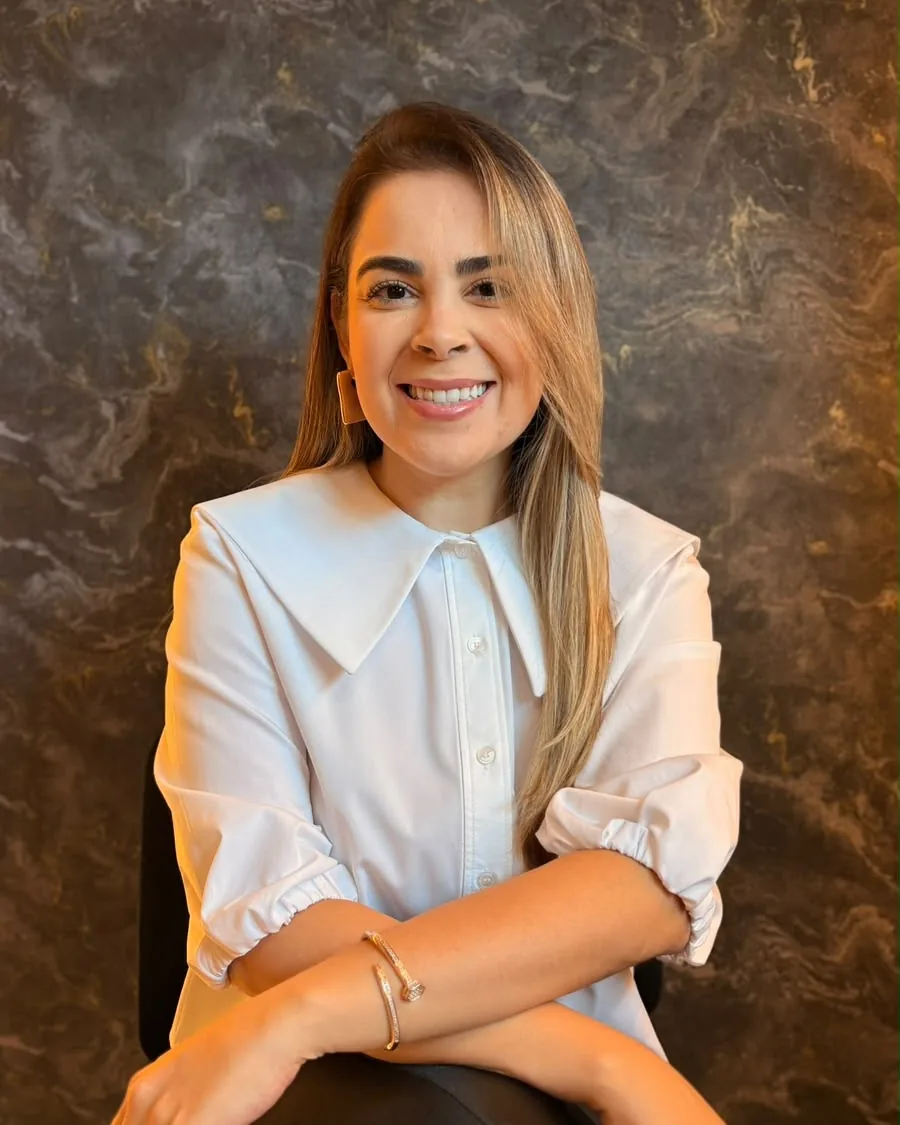 Dra. Fabiana Negreiros sorrindo, de camisa branca, sentada diante de uma parede de mármore escuro